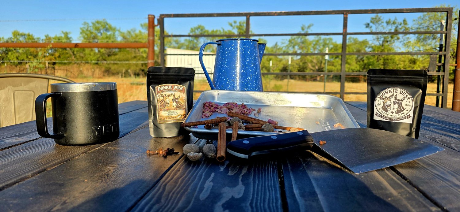 A Yeti mug, two Donkie Dust pouches, the coffee pot, and fresh-cut cinnamon, clove, and nutmeg laid out on a ranch table at sunrise