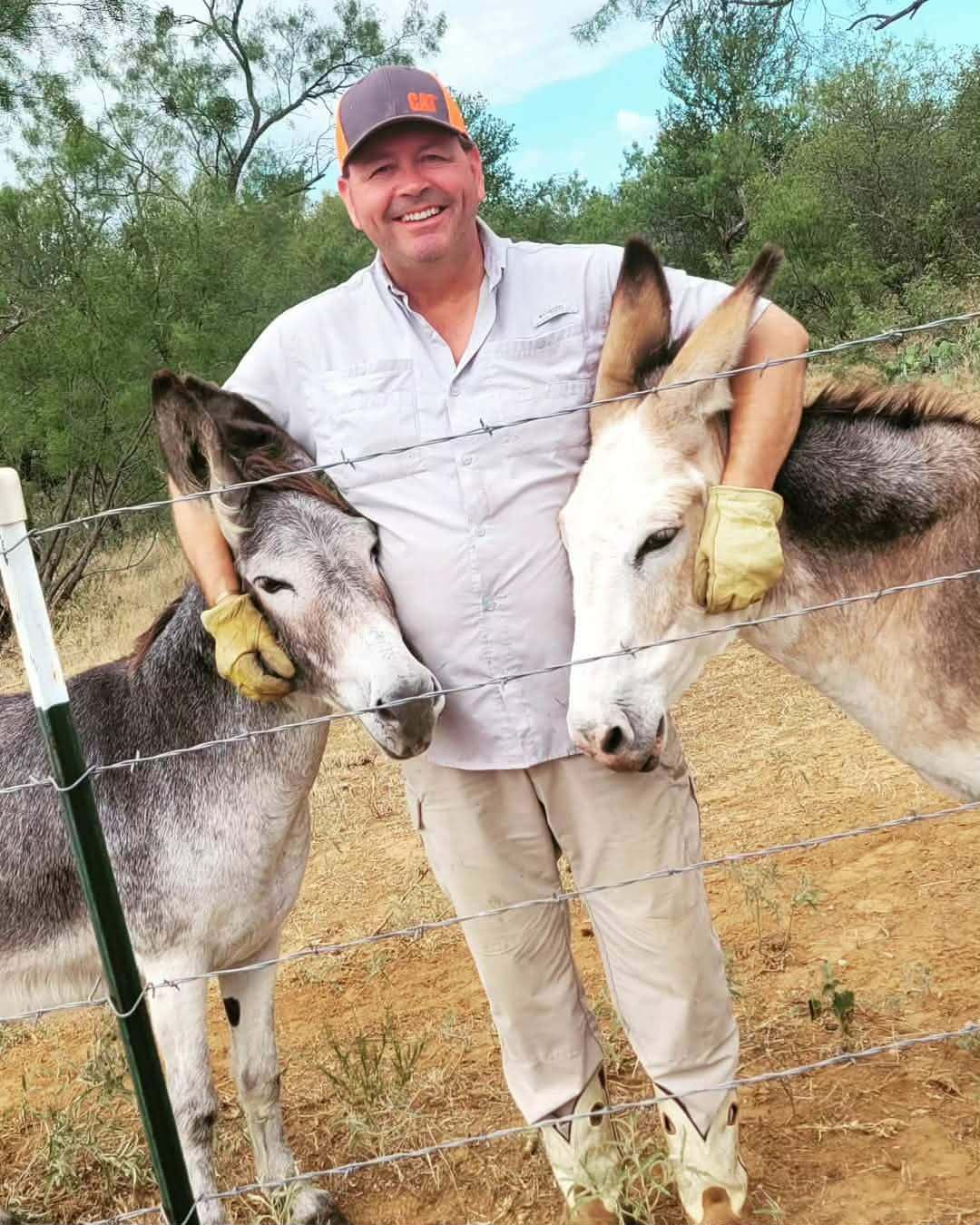 A man leaning in between Thelma and Lulu, arms around both donkeys, at the ranch fence